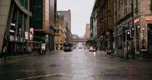 A rainy urban street scene in Glasgow, Scotland, viewed from street level. Wet asphalt reflects headlights and traffic signals, with historic stone buildings and modern storefronts lining both sides. A city bus travels down the road, pedestrians walk along the pavements, and a pedestrian bridge spans the street in the distance. Overcast, atmospheric weather.