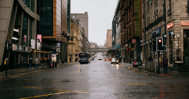 A rainy urban street scene in Glasgow, Scotland, viewed from street level. Wet asphalt reflects headlights and traffic signals, with historic stone buildings and modern storefronts lining both sides. A city bus travels down the road, pedestrians walk along the pavements, and a pedestrian bridge spans the street in the distance. Overcast, atmospheric weather.