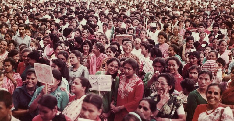 The photo shows a large crowd of standing women stretching far back. They are all facing in the same direction and some hold plackards. Many of the women are smiling and chatting to each other. The scene looks convivial.