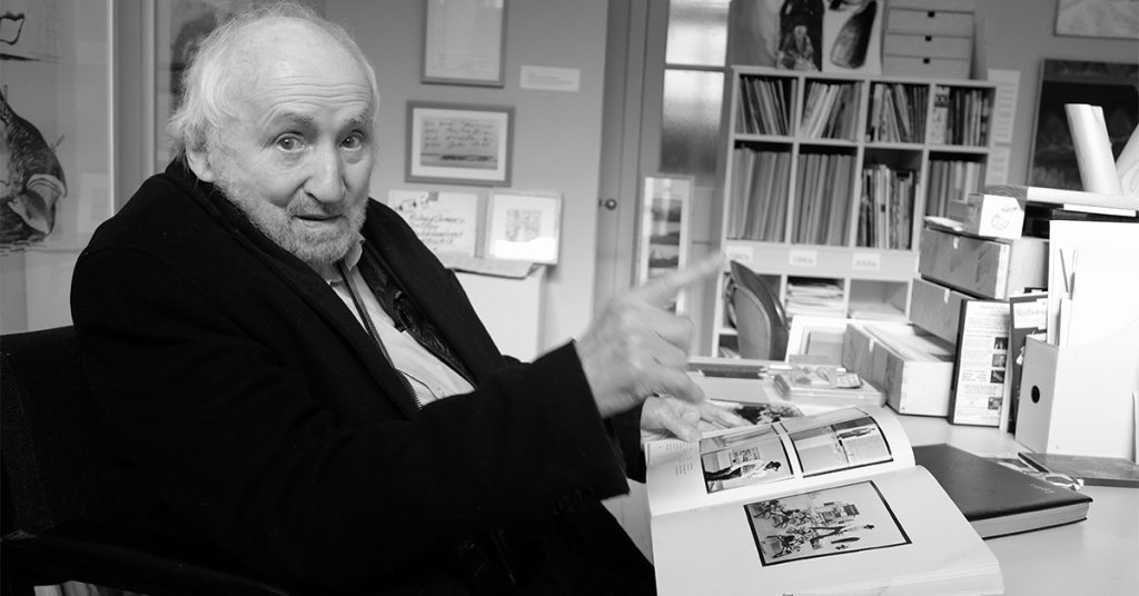 Black-and-white photograph of an elderly man with white hair and a beard sitting at a desk, looking toward the camera while holding open a large illustrated book in an art-filled studio or office.